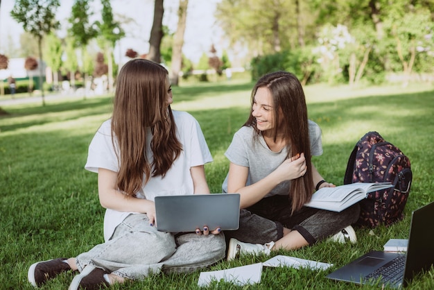 Two female students are sitting in the park on the grass with books and laptops, studying and preparing for exams. Distance education. Soft selective focus.