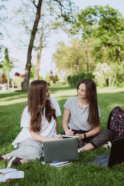 Two female students are sitting in the park on the grass with\
books and laptops, studying and preparing for exams. distance\
education. soft selective focus.