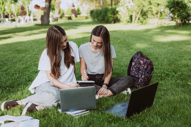 Two female students are sitting in the park on the grass with\
books and laptops, studying and preparing for exams. distance\
education. soft selective focus.