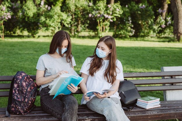 Two female students wearing protective medical masks prepare for exams on a campus bench. Distance education. Soft selective focus.