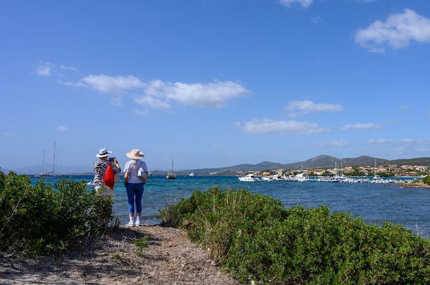 Two female tourists in hats rear view stand on the shore and look at the sea in the background