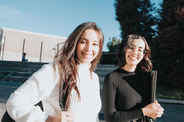 Two female young university colleagues walking smiling to the camera on campus and chatting on a sunny day. Holding backpack and laptop.Study and learn concepts. Confident and happy. Sunny day.