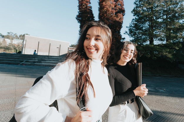 Two female young university colleagues walking smiling to the camera on campus and chatting on a sunny day. Holding backpack and laptop.Study and learn concepts. Confident and happy. Sunny day.