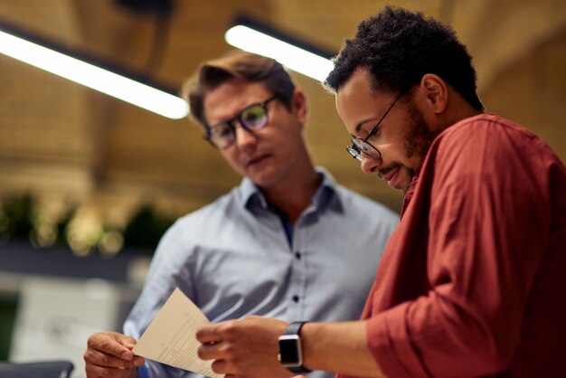 Two focused diverse business colleagues analyzing document and discussing something while standing in coworking space, working together. Office life, teamwork and business concept