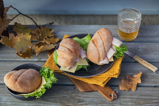 Two fresh ciabatta baguette sandwiches with ham, cheese, lettuce and a glass of beer on rustic wooden background. Outdoor autumn picnic lifestyle.