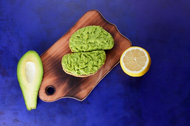 Two fresh green avocado toasts on a wooden board Toasts half an avocado and a yellow lemon in a peel on a blue background Closeup