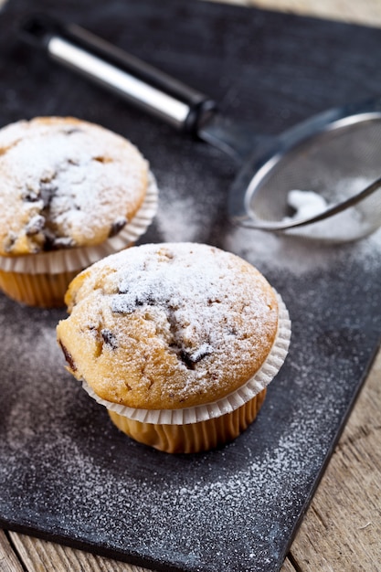 Two fresh homemade muffins with sugar powder and metal strainer closeup on black board.