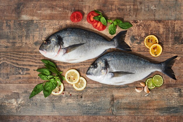 Two fresh uncooked dorado with lemons, herbs, tomatoes and spices on wooden table. Top view