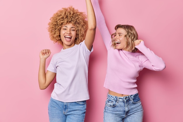 Two friendly girls have fun dance carefree shake arms chill together have glad expressions foolish around dressed in casual clothes isolated over pink background People and entertainment concept