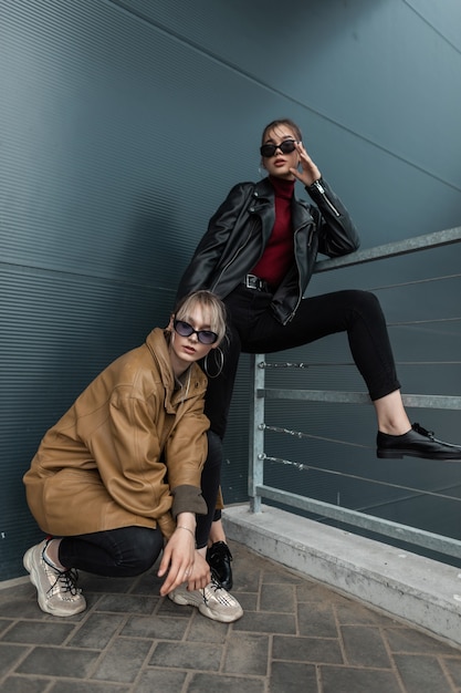 Two friends beautiful young women in fashionable clothes with leather twist and jeans posing on the street