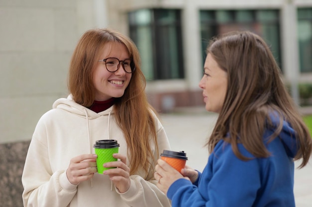 Two friends girls young women students chat talk with each other and drink coffee outdoors at campus