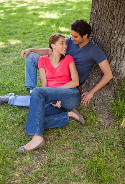 Two friends looking at each other while sitting together against a tree