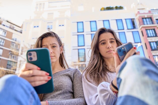 Two friends sitting on a city bench bored using their cell phone