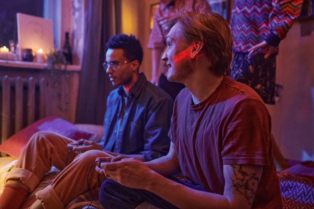 Two friends sitting on the floor in dark room and playing video games during party