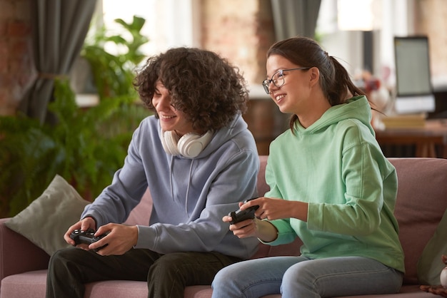 Two friends sitting on sofa using joysticks to play video games in team in the room at home