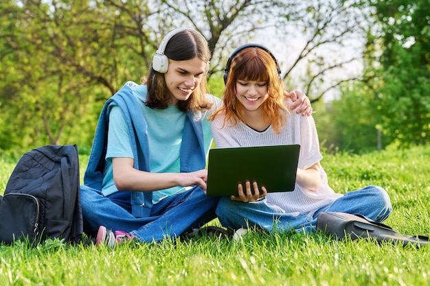 Two friends students guy and girl in headphones looking at laptop sitting on grass