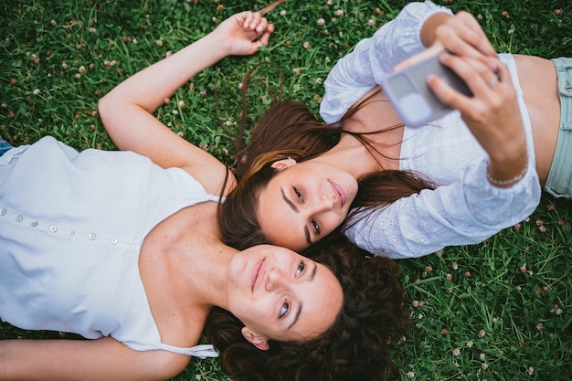 Two friends taking a selfie photo while they are laying down on the grass in a park