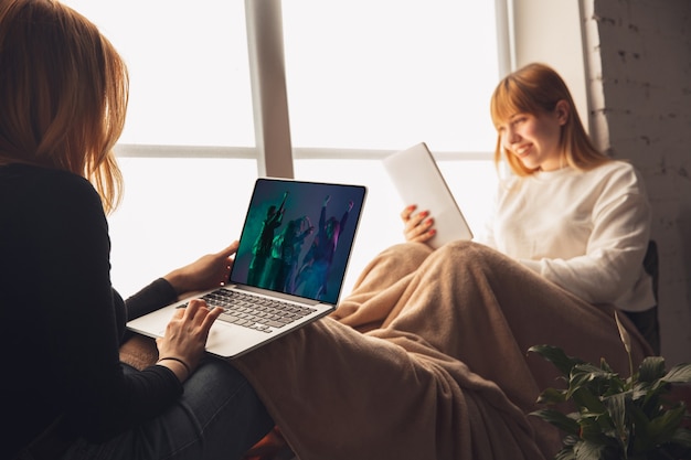 Photo two friends watching online party, tv or cinema together with devices. online entertainment, communication during quarantine. prevention of spreading of covid-19, coronavirus. pretty emotional girls.