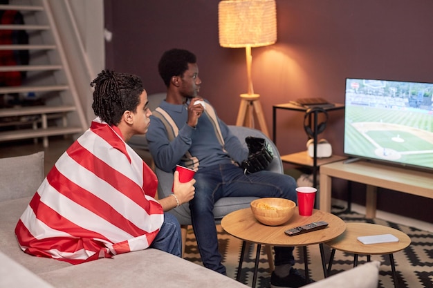 Two friends watching sports match on TV in living room and drinking beer