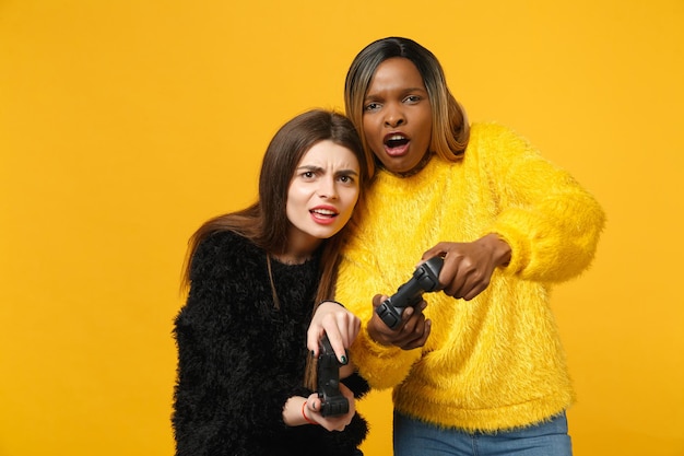 Two fun young women friends european and african american in black yellow clothes hold joystick isolated on bright orange wall background, studio portrait. People lifestyle concept. Mock up copy space