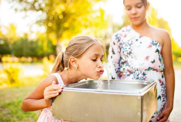 Two funny cheerful wonderful sisters drink cool fresh water from a small fountain in a summer warm sunny park on a long-awaited vacation