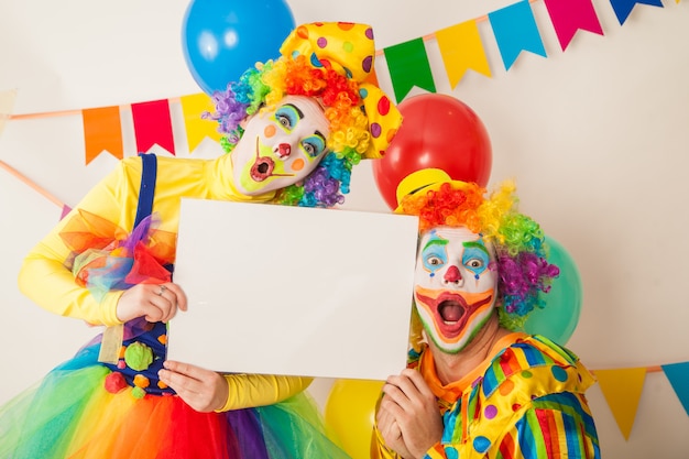 Two funny clowns at the holiday hold a white sign for inscriptions