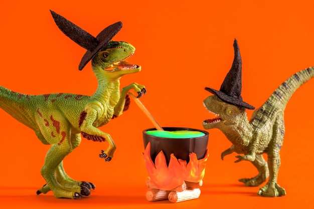 Фото Два забавных динозавра в шляпах ведьм варят зелье в кастрюле на оранжевом фоне. открытка на хэллоуин