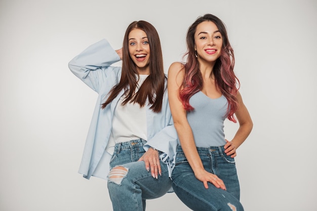 Two funny fashion beautiful cheerful girls girlfriends with smile in trendy stylish denim clothes have fun and pose on a white background in the studio