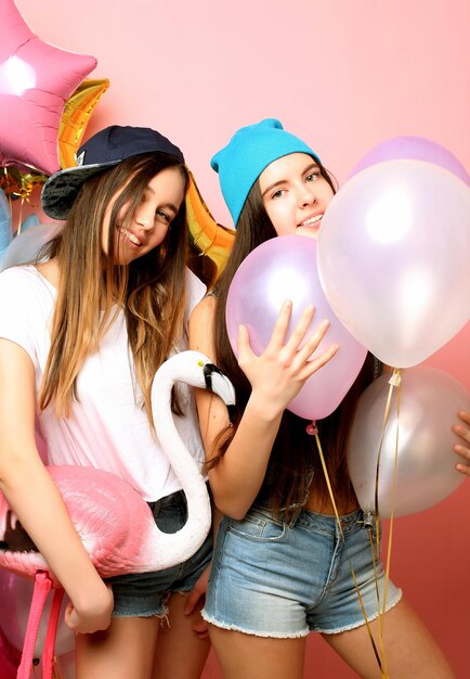 Two funny friends make grimace and have fun Two emotional girls hold balloons and posing against pink wall