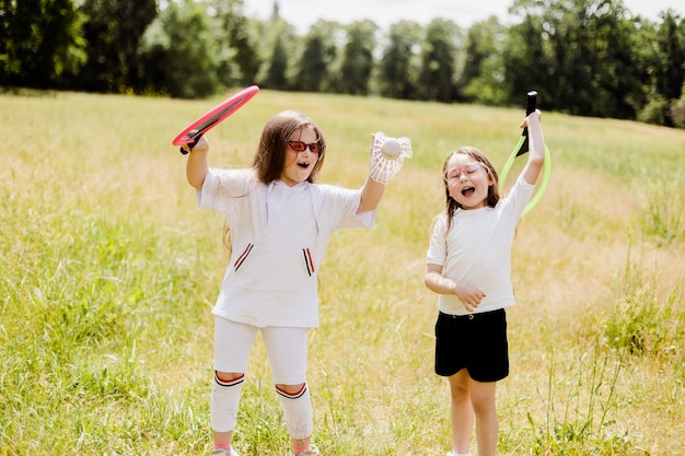 Two funny little girls best friends or sisters with badminton rackets playing in summer park