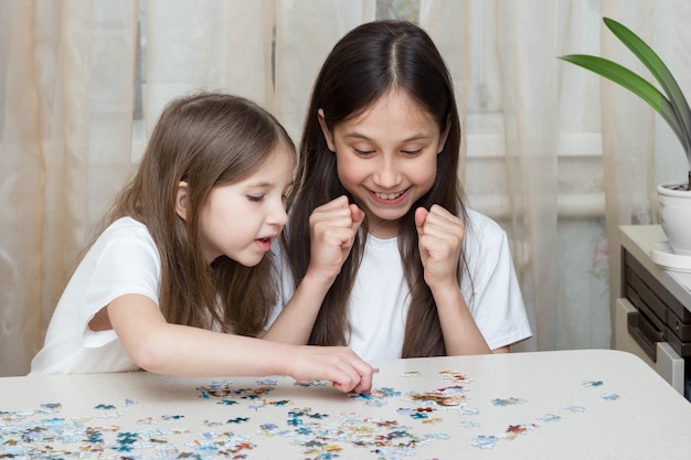 2人の面白い小さな女の子の姉妹がパズルを集めている窓のそばのテーブルで一緒に遊んでいます