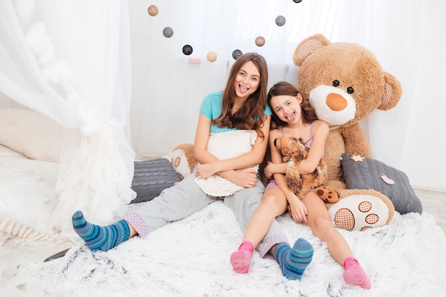 Two funny lovely sisters sitting and showing tongues in children room