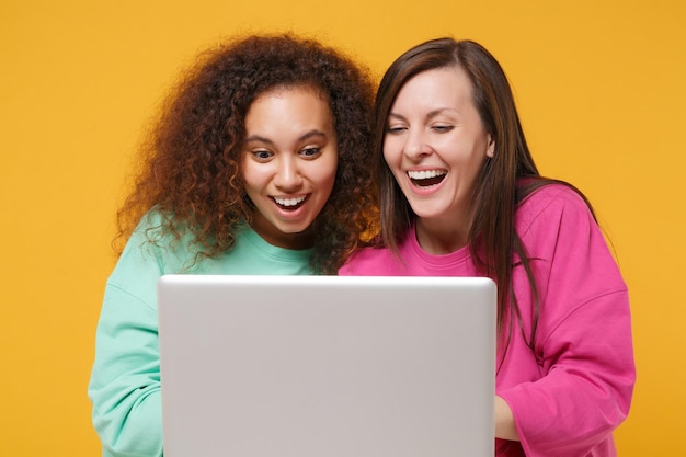 ピンクの緑の服を着た2人の面白い女性の友人のヨーロッパ人とアフリカ系アメリカ人の女の子が黄色いオレンジ色の背景に分離してポーズをとっています。人々のライフスタイルのコンセプト。コピースペースをモックアップします。ラップトップPCコンピューターを保持します。