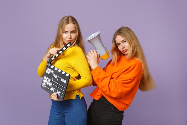 Two funny young blonde twins sisters girls holding classic black film making clapperboard, scream on megaphone isolated on violet blue wall . People family lifestyle concept. .