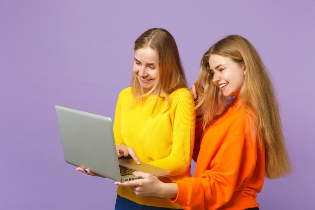 Две смешные молодые блондинки сестры-близнецы девушки в яркой красочной одежде держатся, используя портативный компьютер, изолированный на пастельной фиолетовой синей стене. Концепция семейного образа жизни людей. .