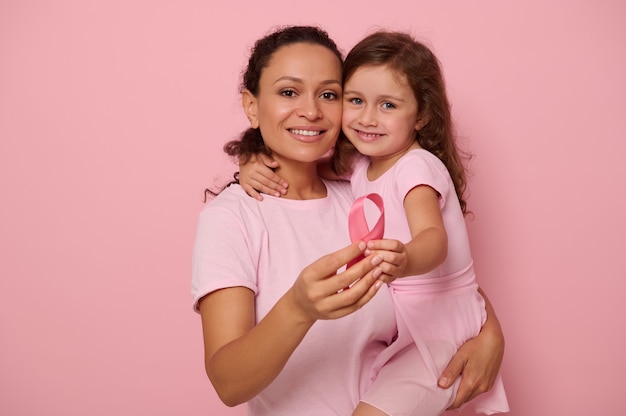 Two generations of women, woman and girl, mother and daughter posing against a pink background, holding a pink ribbon, showing support and solidarity to patients and cancer survivors. Medical concepts