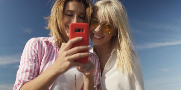 Two girl friends taking photo with smartphone on empty beach during autumn day