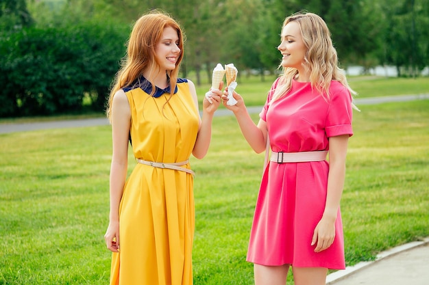 Two girlfriends beautiful young ginger redhead woman in a yellow dress and blonde female person in a pink dress eating vanilla ice cream in a waffle cone in the summer park