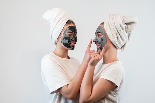 Foto due amiche o sorelle in una maglietta bianca e un asciugamano sulla testa applicando maschera di argilla isolata su bianco concetto di trattamento anti invecchiamento per la cura della pelle