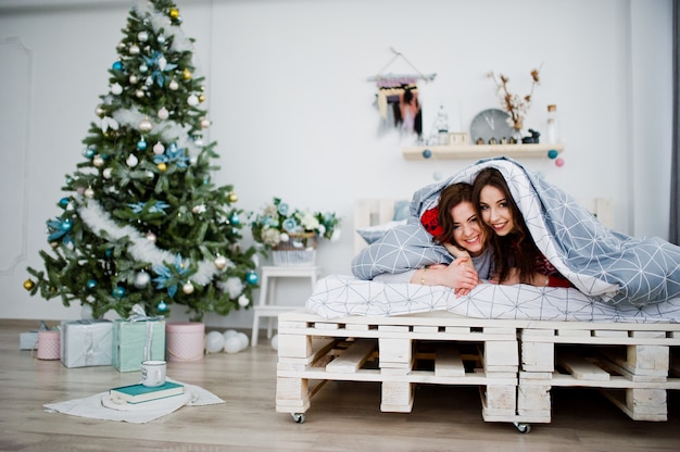 Two girlfriends wear on winter sweaters having fun on bed at room with chrismas decorations.