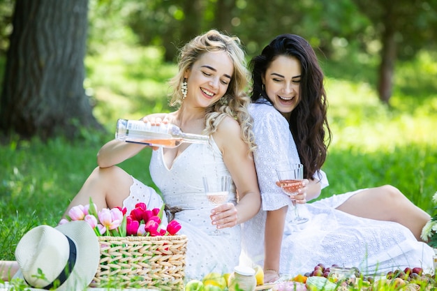 Two girls are resting in park sitting on a picnic blanket with fruits and wine