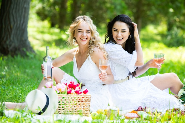 Two girls are resting in park sitting on a picnic blanket with fruits and wine