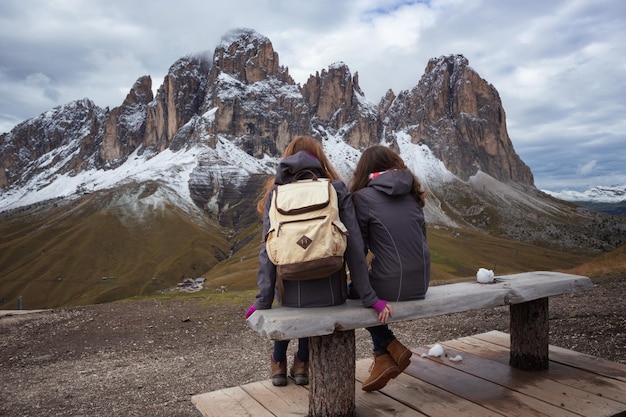 Two girls are sitting on a bench and view of Sassolungo Langkofel. Dolomites, Italy.