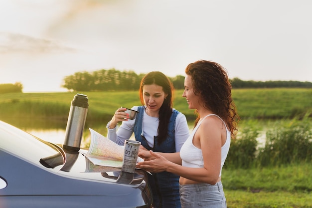 2人の女の子が道路の脇に停車している車で道路を移動し、魔法瓶休暇のコンセプトからコーヒーを飲みながら地図を見る