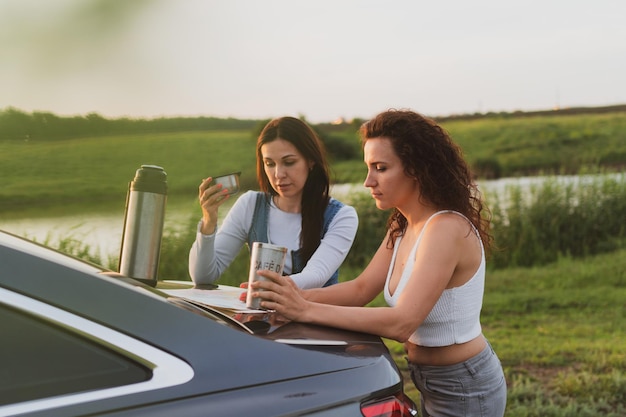 Две девушки едут по дорогам на машине, остановились на обочине и смотрят на карту, пьют кофе из термоса Концепция отпуска