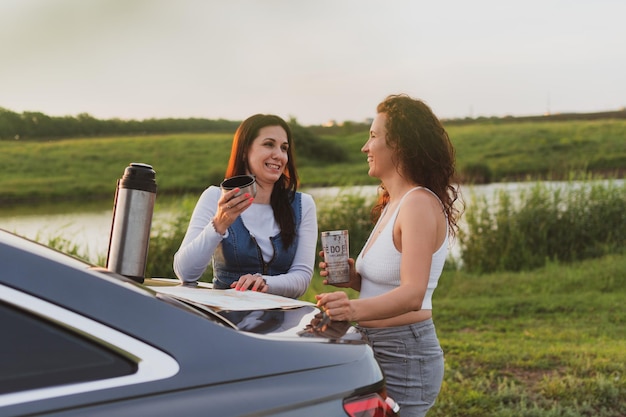 Two girls are traveling on the roads by car stopped at the side of the road and look at the map drinking coffee from a thermos Vacation concept