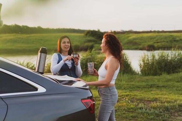 Two girls are traveling on the roads by car stopped at the side\
of the road and look at the map drinking coffee from a thermos\
vacation concept