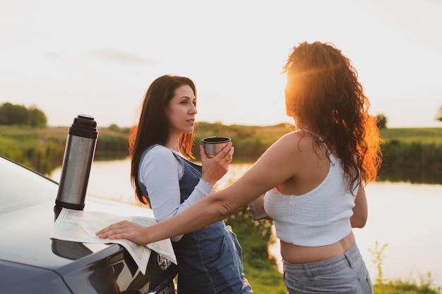 Two girls are traveling on the roads by car stopped at the side of the road and look at the map drinking coffee from a thermos Vacation concept