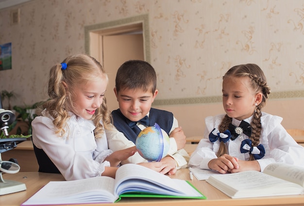 Две девочки и мальчик в школе за партой изучают географию, наблюдая за земным шаром
