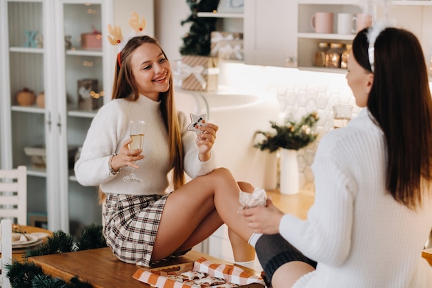 Two girls in a cozy home environment in the kitchen with champagne in their hands for Christmas. Smiling girls drink champagne on a festive evening